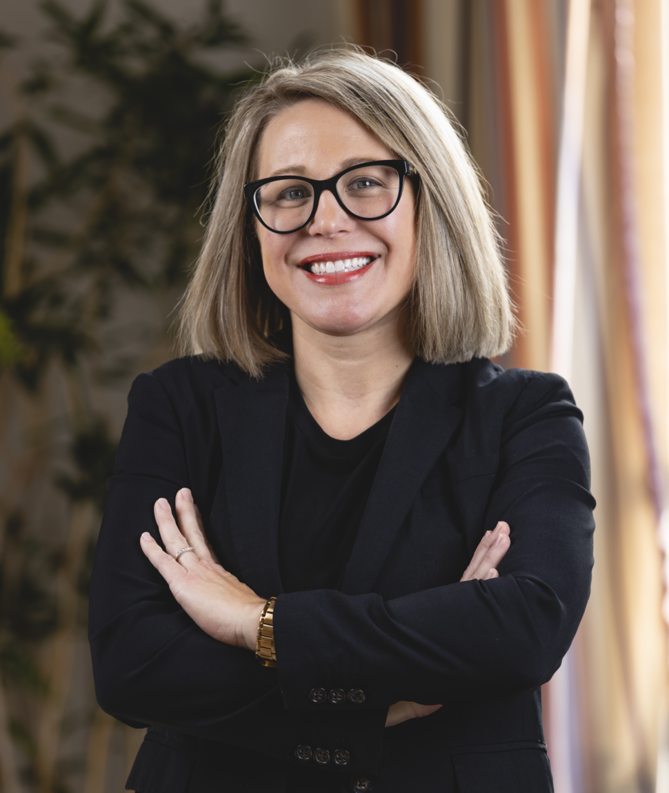 Woman (Jennifer Pegher) with blonde hair and glasses wears a suit and smiles at the camera