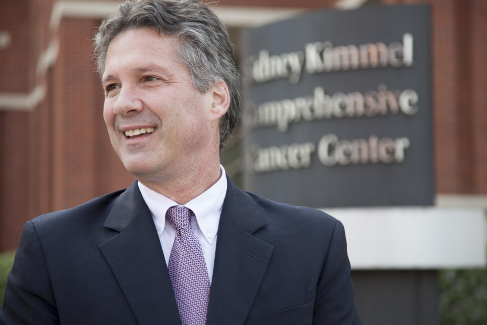 Man (William Nelson) with grey hair wears a suit and tie and smiles in front of a blurry sign that reads: Sidney Kimmel Comprehensive Cancer Center