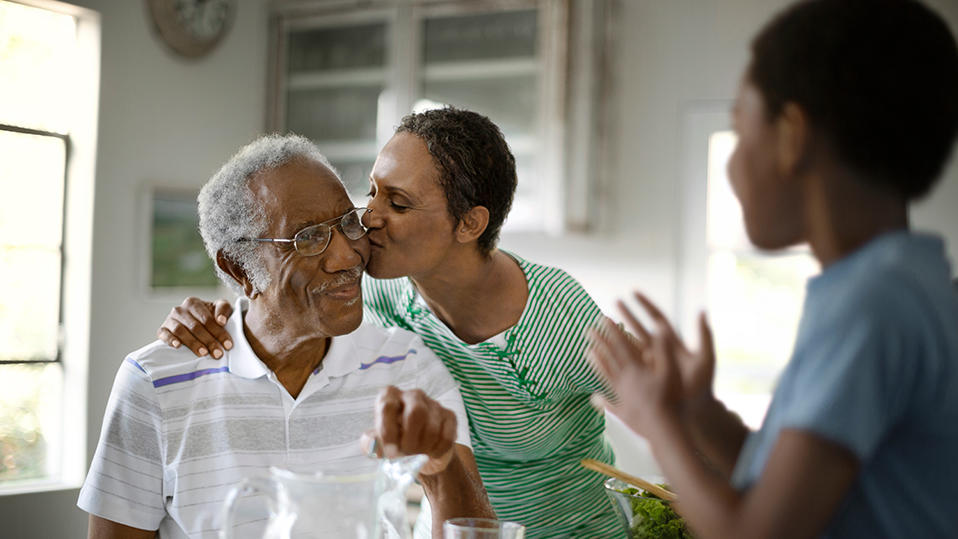 A warm family moment in a kitchen setting. An elderly, man with glasses and short, white hair smiles as a woman with dark, short hair kisses him on the cheek, her arm around his shoulders. A young person, seen from behind is blurred in the foreground. A pitcher of lemonade and a bowl of greens are on the table.